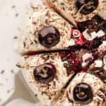 Overhead view of a white forest cake decorated with swirls of frosting, grated chocolate and white chocolate, cherry compote and fresh cherries cut into slices with text overlay.