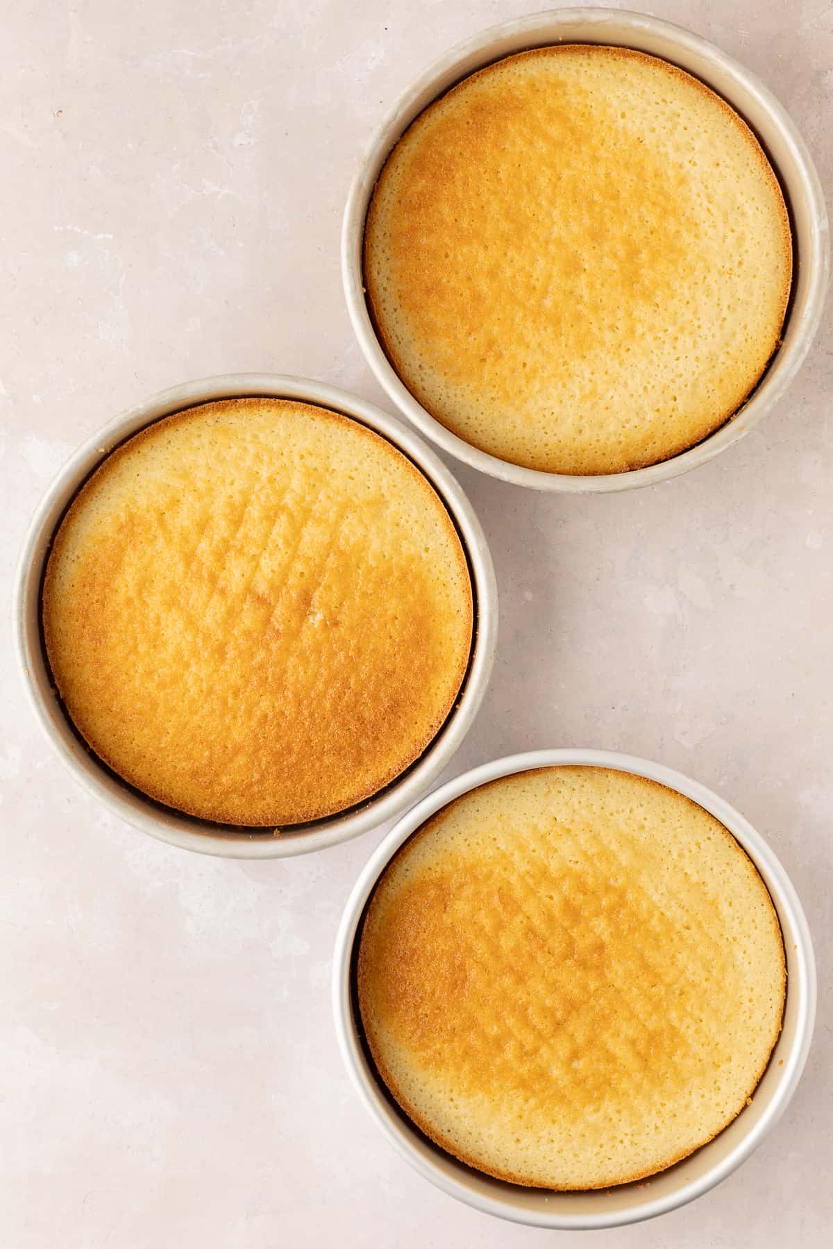 Three baked white cakes in their pans over a light pink surface.