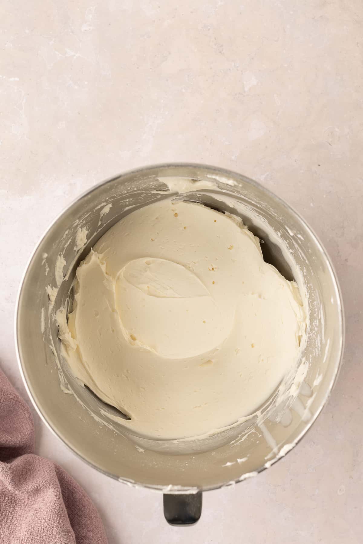 Whipped cream cheese frosting in a silver mixing bowl over a light pink background.