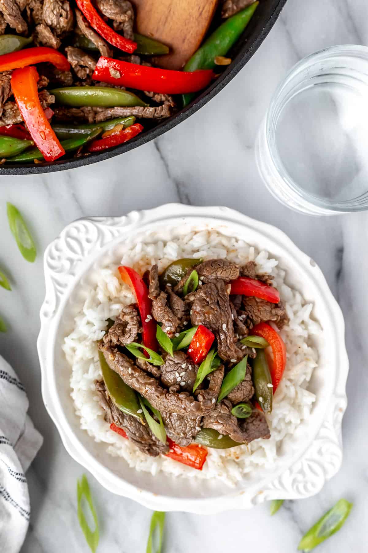 Overhead view of ginger beef stir fry with sugar snap peas and red peppers in a white bowl with rice with a skillet and glass of water partially showing in the background.