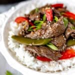 Close up of ginger beef stir fry with sugar snap peas and red peppers in a white bowl with rice.