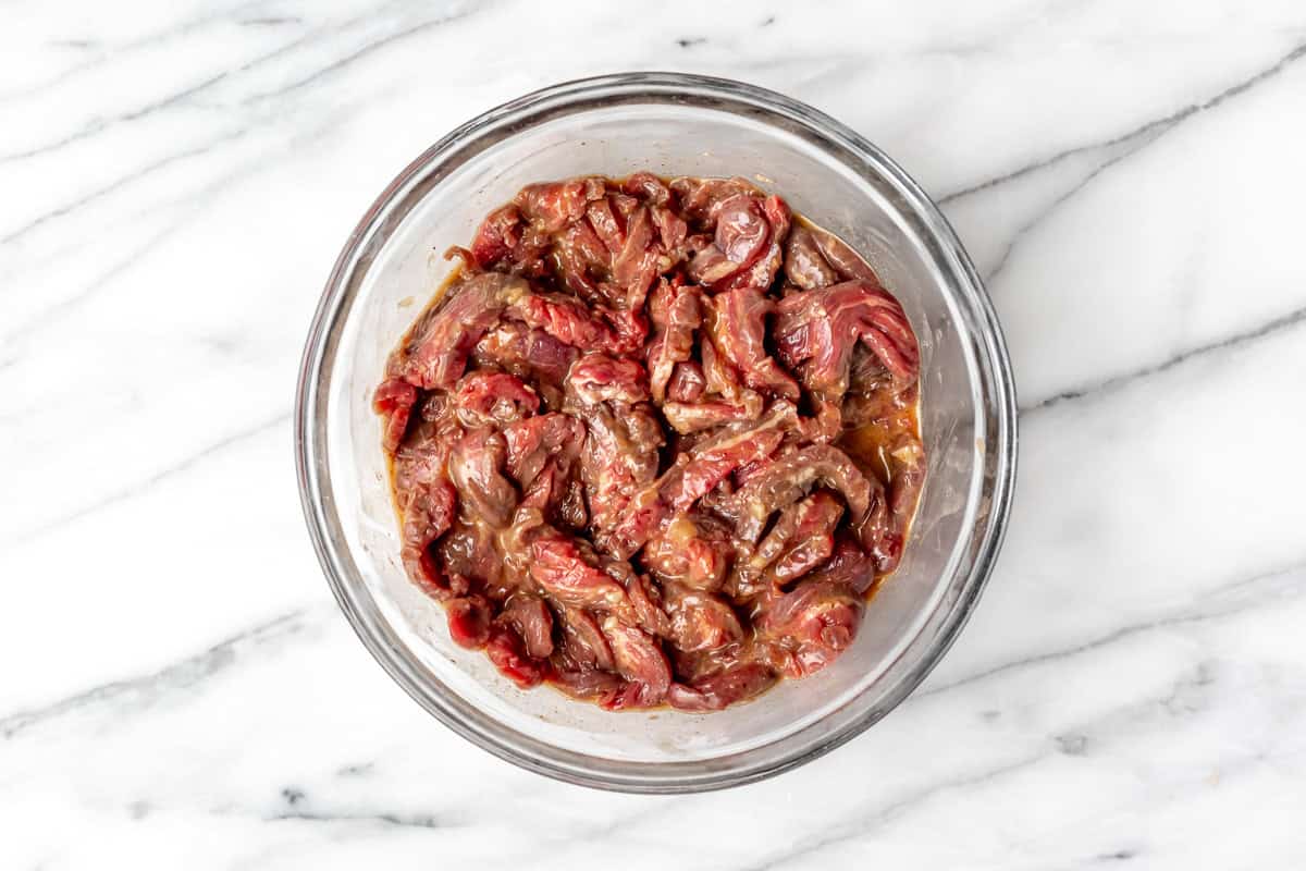 Thin strips of beef marinating in a glass bowl over a marble background.