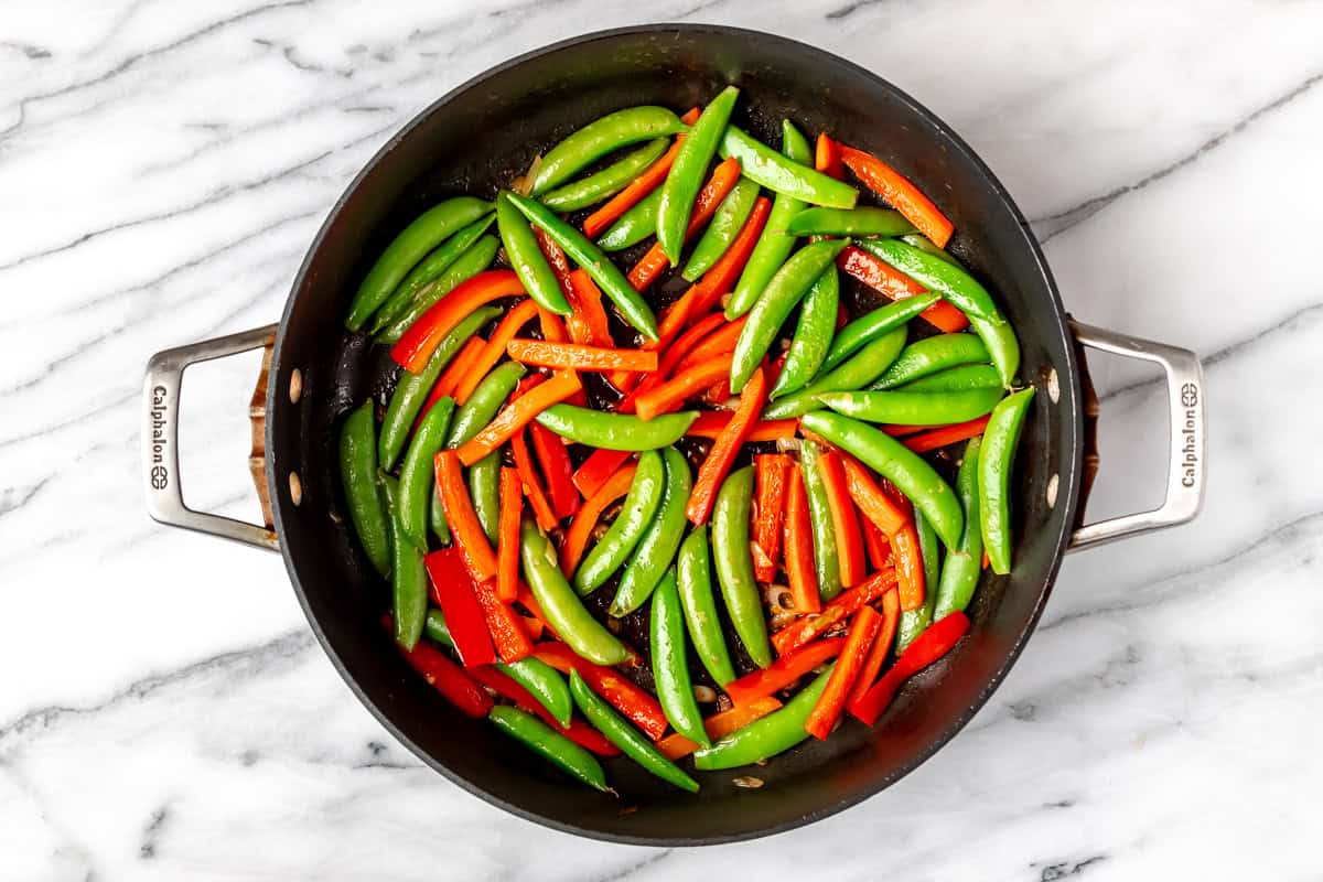 Sugar snap peas and red bell pepper strips in a black skillet over a marble background.