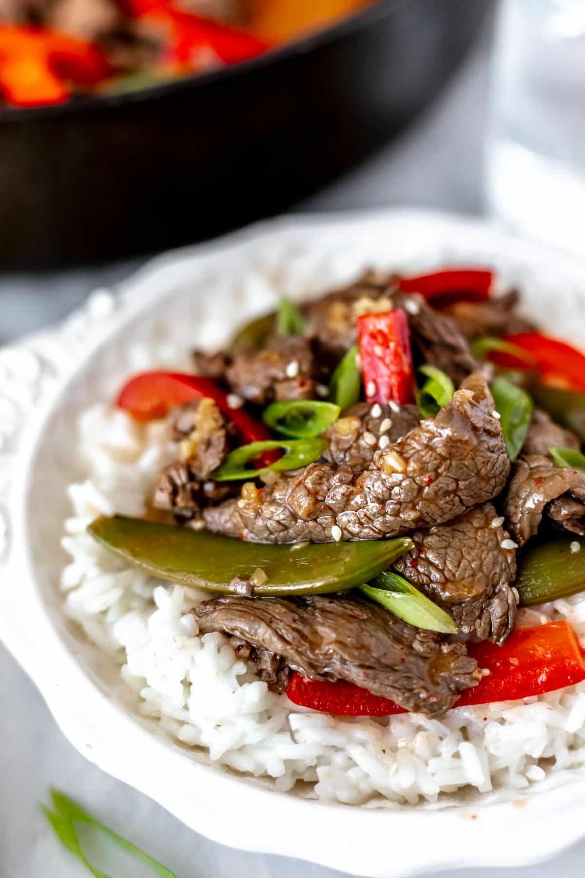 Close up of ginger beef stir fry with sugar snap peas and red peppers in a white bowl with rice with a skillet partially showing in the background.