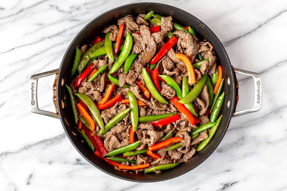 Ginger beef, sugar snap peas and red bell peppers in a black skillet over a marble background.