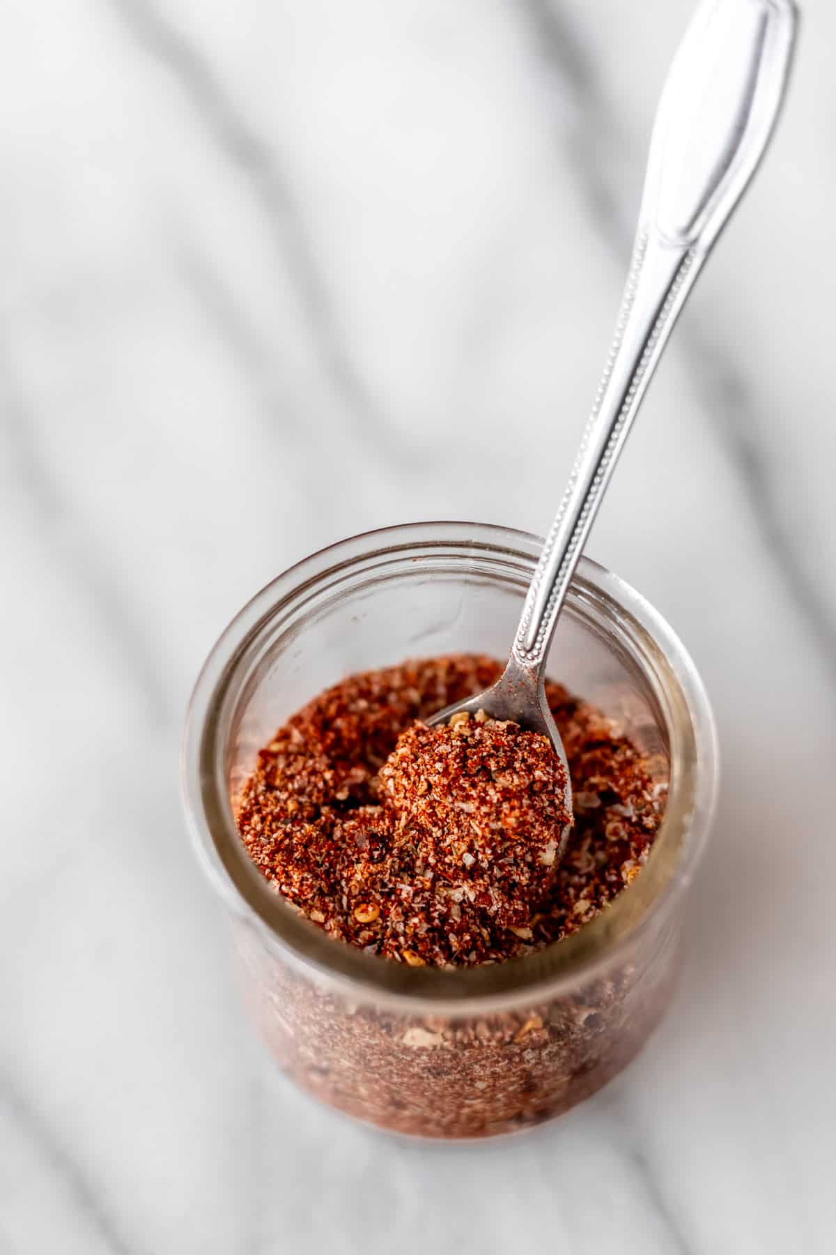 Homemade Montreal steak seasoning in a small glass jar with a small spoon in it on a marble background.