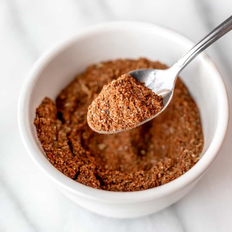 A spoonful of homemade Old Bay Seasoning being held up over a small white bowl filled with more seasoning.