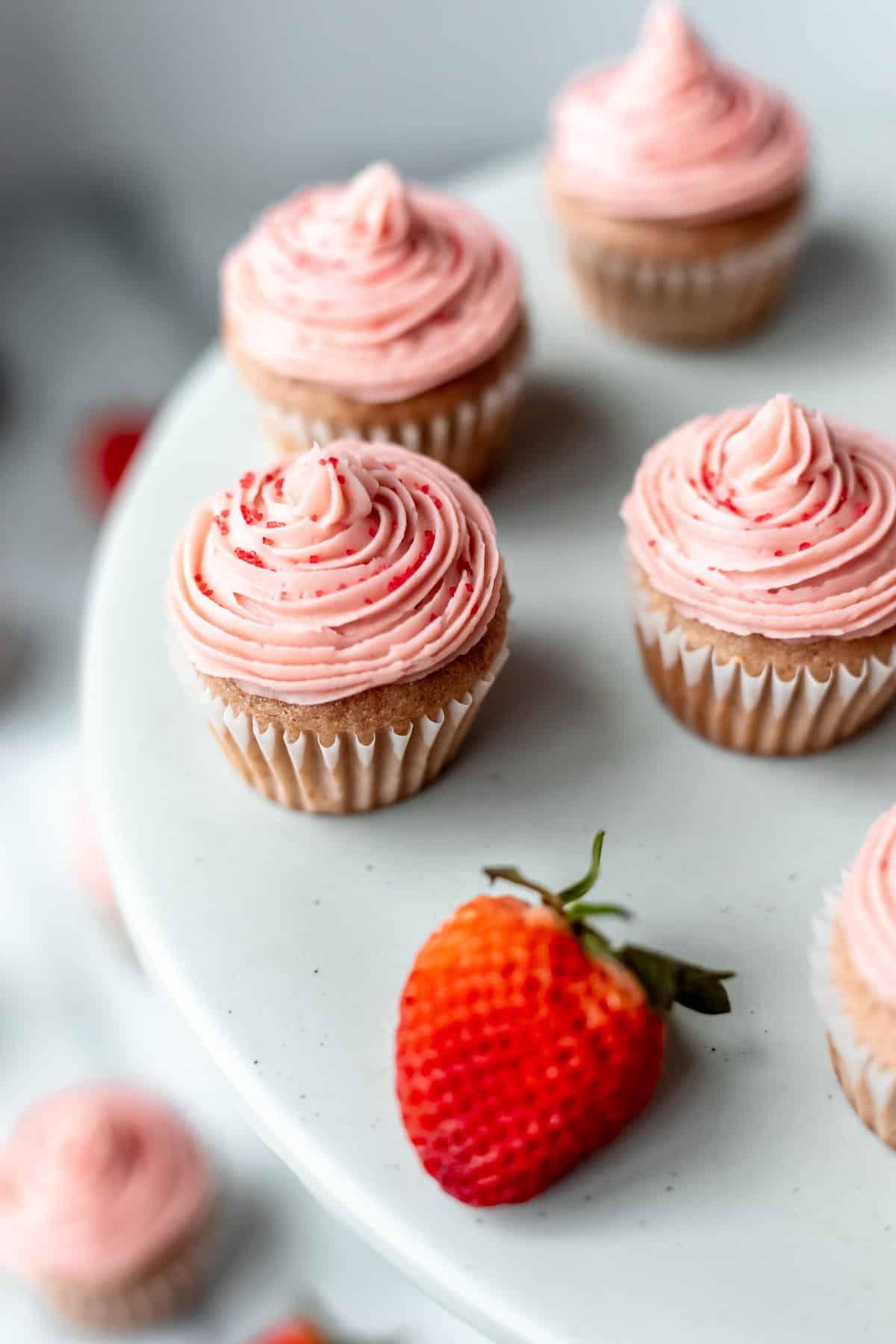 Mini strawberry cupcakes topped with strawberry buttercream frosting on a gray cake stand with strawberries.