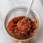 Homemade Montreal steak seasoning in a small glass jar with a small spoon in it on a marble background with text overlay.