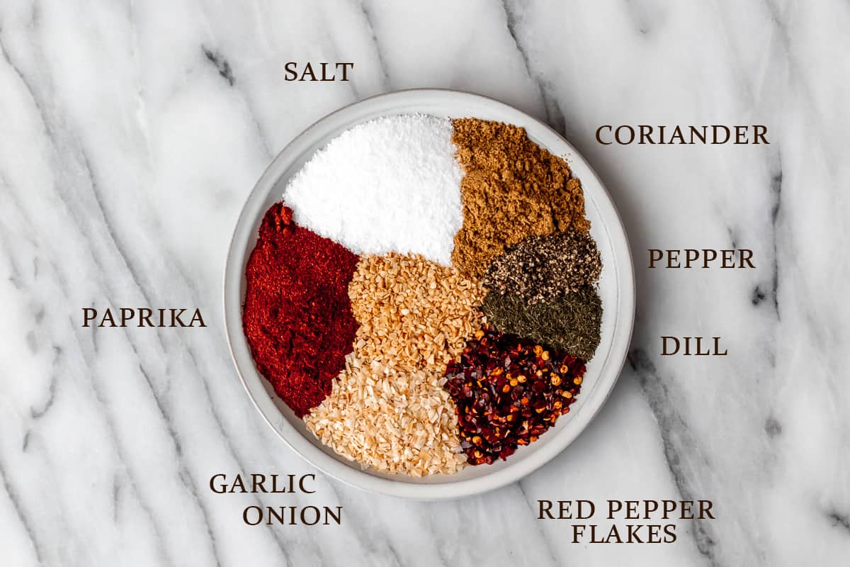 Ingredients needed to make homemade Montreal steak seasoning on a white plate on a marble background with text overlay.
