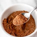 A spoonful of homemade Old Bay Seasoning being held up over a small white bowl filled with more seasoning with text overlay.