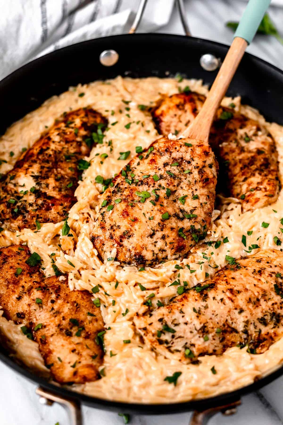 Parmesan Chicken Orzo in a skillet with a wood spoon lifting up a piece of chicken.