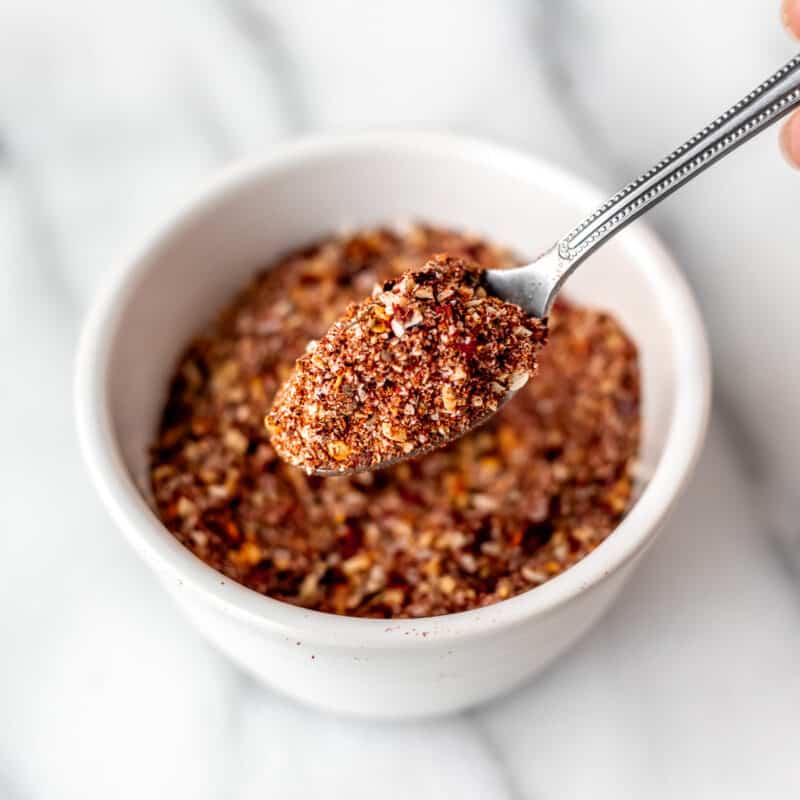 A spoonful of Montreal steak seasoning being held up in front of a small white bowl of more seasonings.