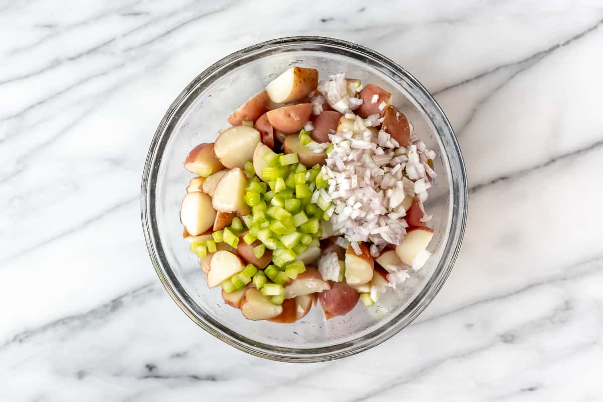 A glass bowl filled with red potatoes, celery and shallots on a marble background.