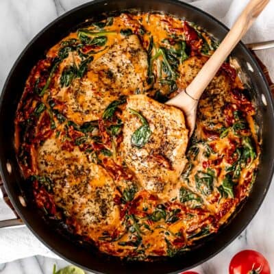 Creamy Tuscan Chicken in a black skillet with one piece being lifted up some with a wood spoon.