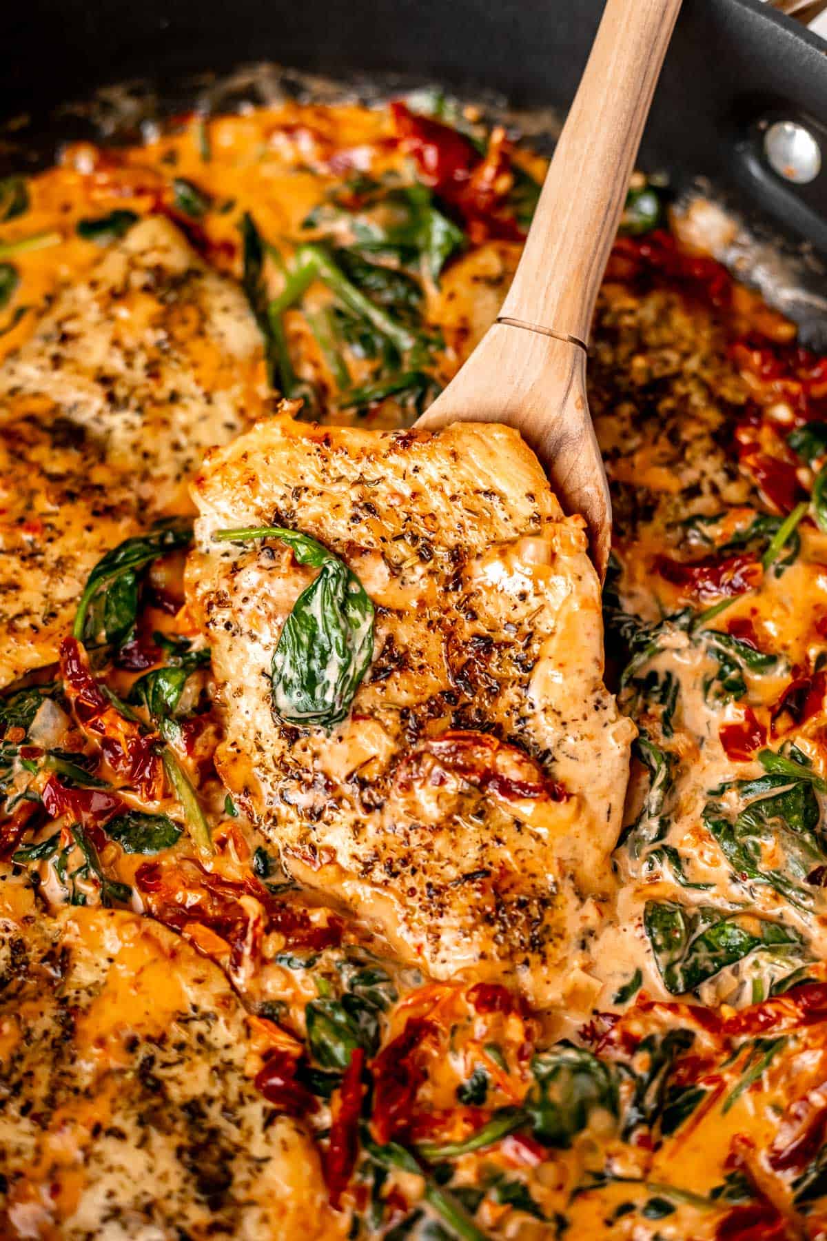 Close up of a Creamy Tuscan Chicken with one piece being lifted up some with a wood spoon.