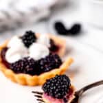 A blackberry tartlet with a bite on a fork in front of it on a small white plate with blackberries, a towel and small white bowl in the background with text overlay.