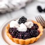 A blackberry tartlet on a white plate topped with whipped cream and a blackberry with other mini tarts partially showing around it with text overlay.