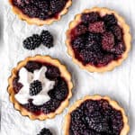 Overhead of 4 blackberry tartlets, one with whipped cream, on a white surface with text overlay.