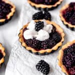 A blackberry tartlet topped with whipped cream and a blackberry with other mini tarts partially showing around it.