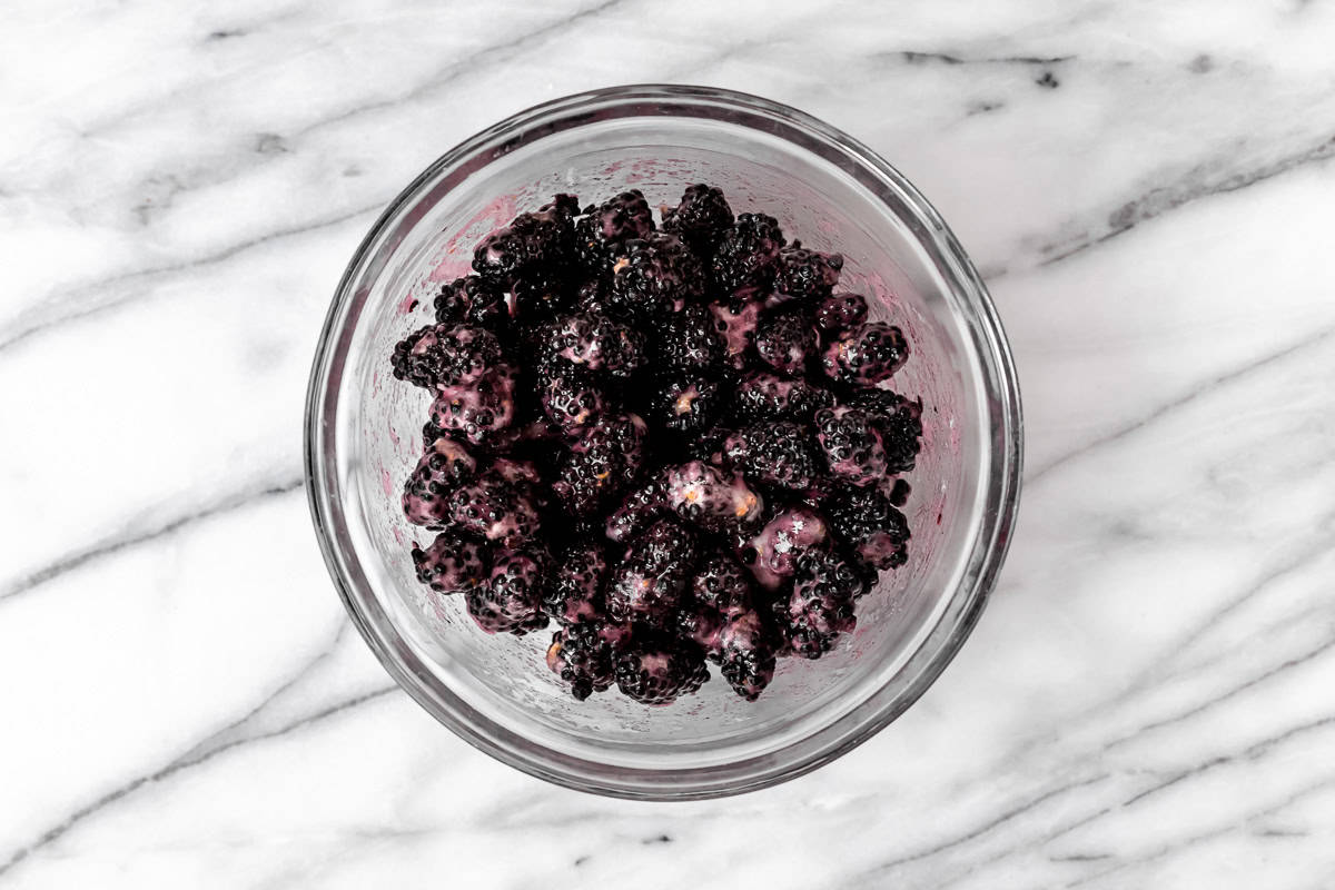 A bowl full of blackberries that have been mixed with sugar, cornstarch and lemon juice on a marble surface.