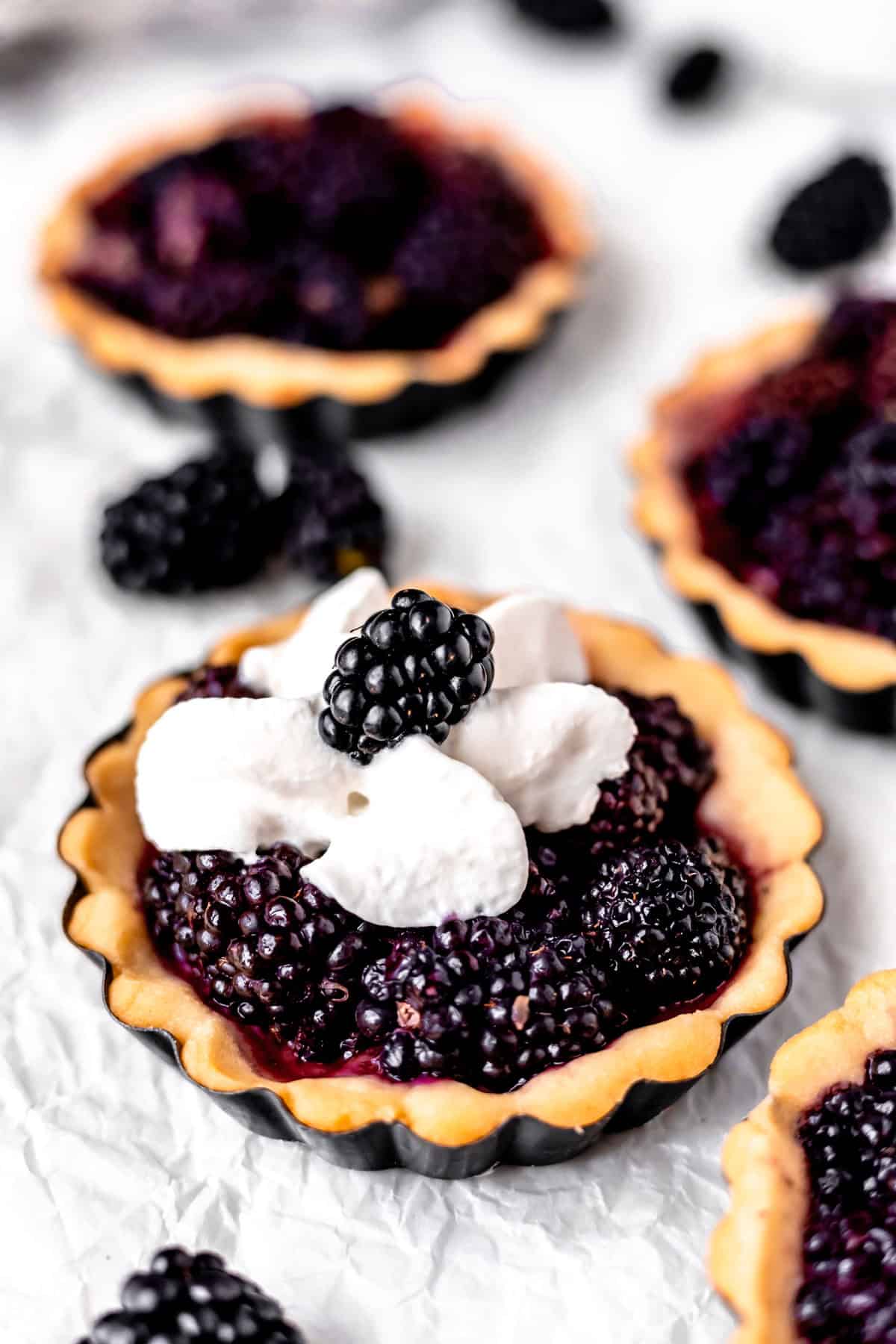 A blackberry tartlet topped with whipped cream and a blackberry with other mini tarts partially showing around it.