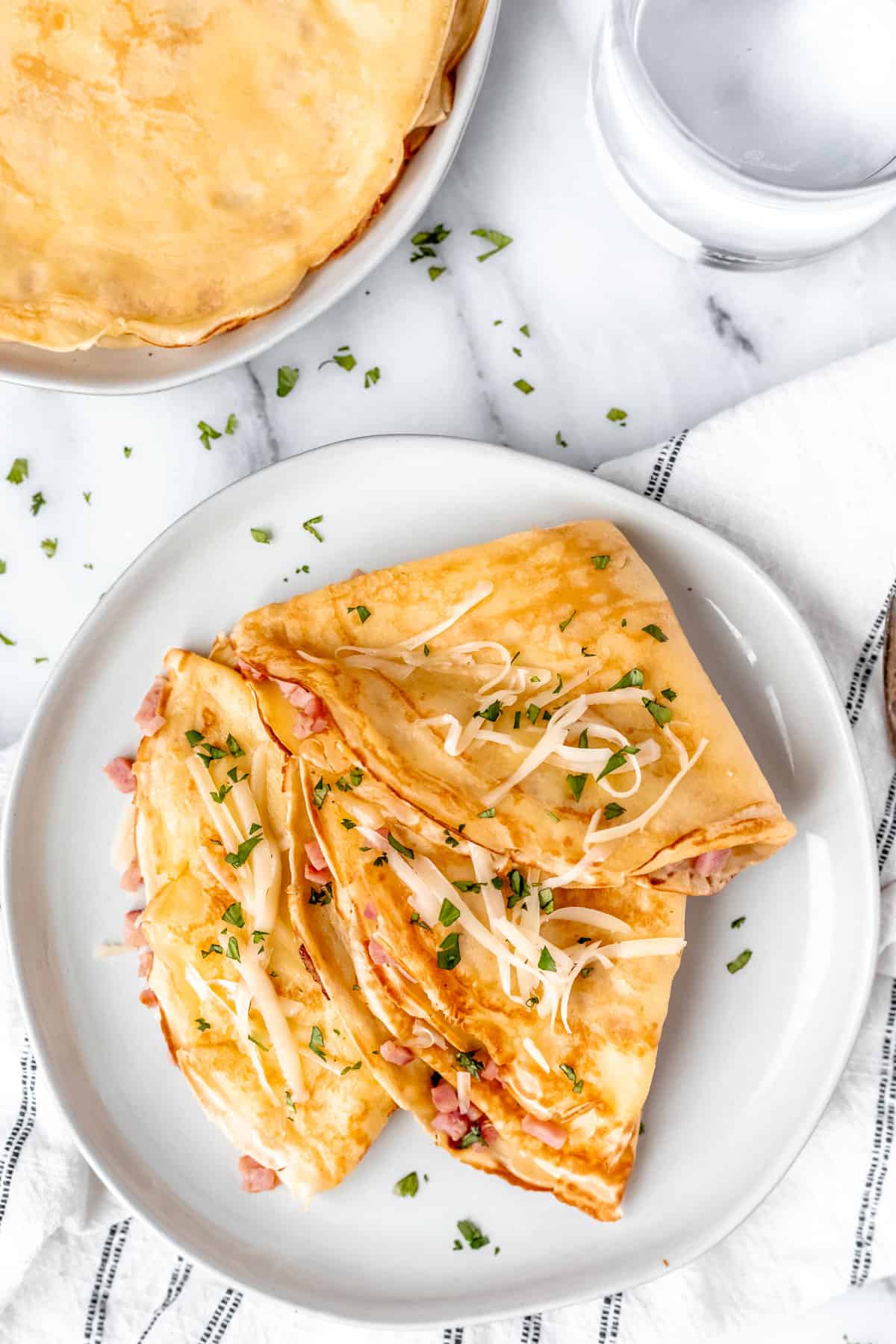 Overhead view of three folded ham and cheese crepes on a white plate toped with cheese and parsley with a plate of crepes and a glass of water partially showing in the background.