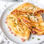 Three folded ham and cheese crepes on a white plate toped with cheese and parsley.