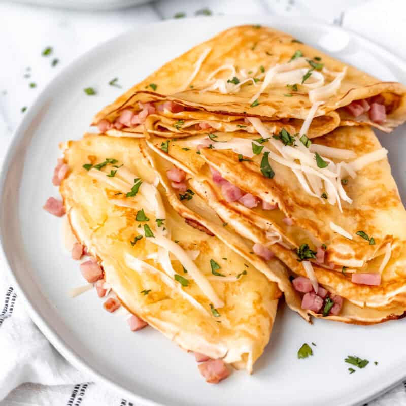 Three folded ham and cheese crepes on a white plate toped with cheese and parsley.