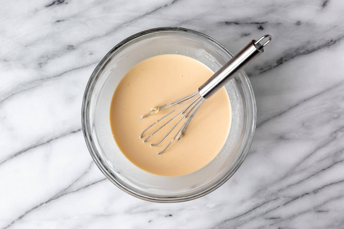 Crepe batter in a glass bowl with a whisk in it over a marble background.