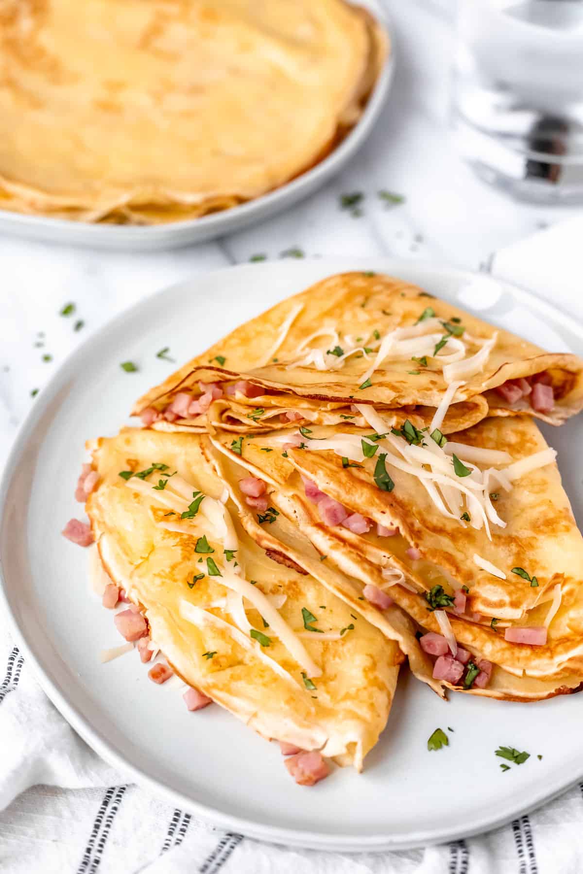 Three folded ham and cheese crepes on a white plate toped with cheese and parsley with a plate of crepes and a glass of water in the background.