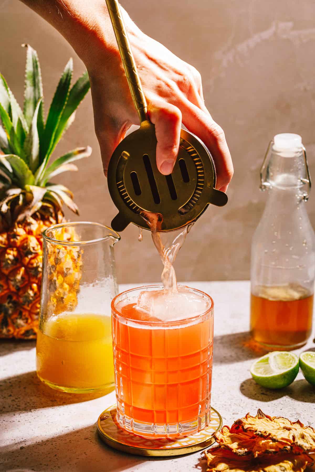 A jungle bird cocktail being strained into a glass with ice with pineapples, limes and juices in the background.