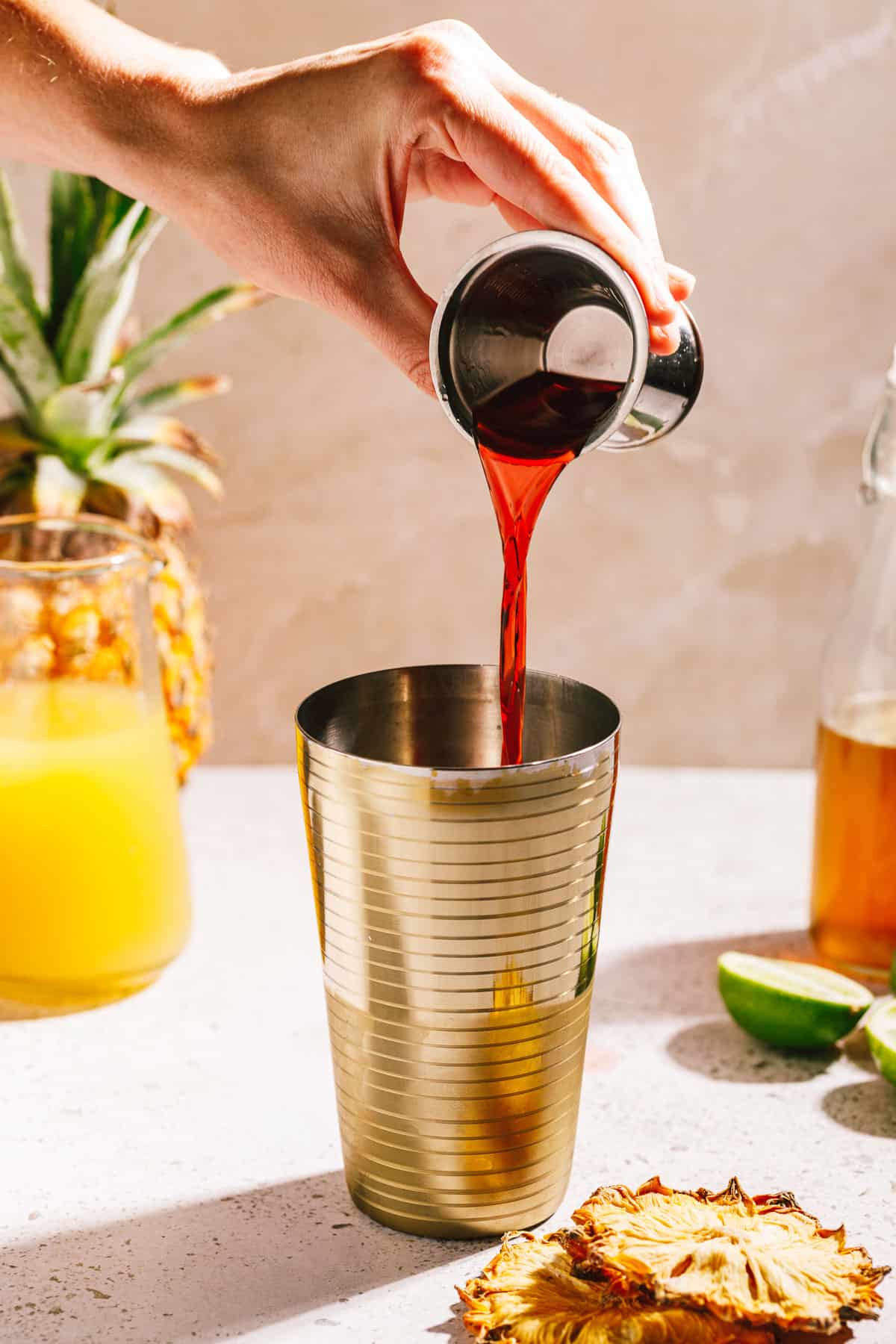 Campari being poured into a cocktail shaker with a pineapple, limes and juices around it.