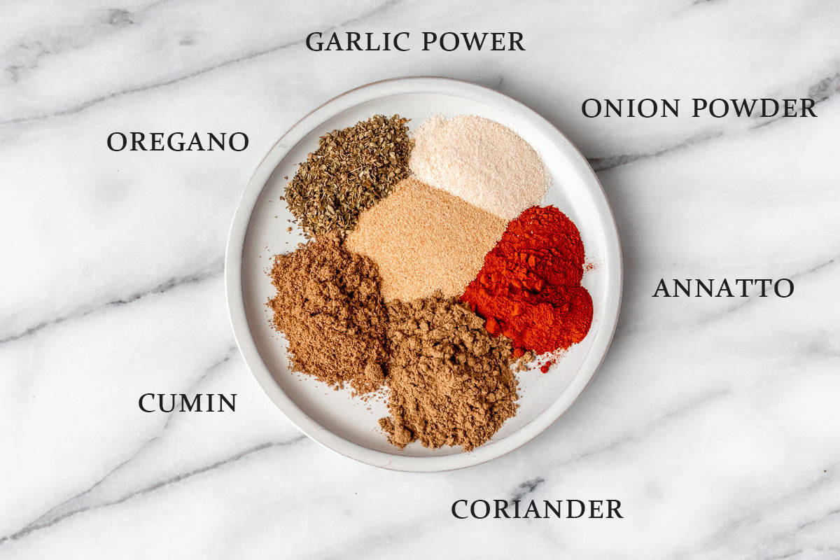 Ingredients needed to make homemade sazon seasoning on a white plate over a marble background with text overlay.