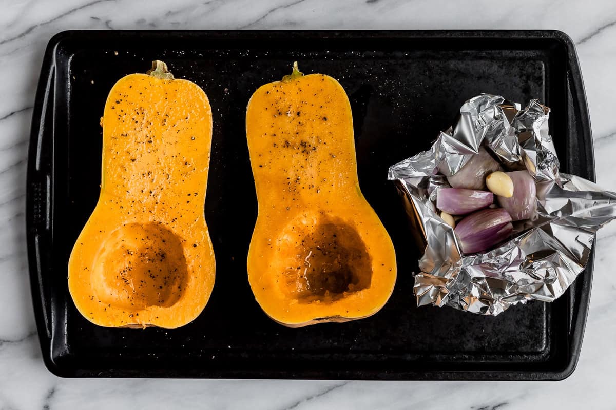 Two halves of butternut squash and shallots in foil on a baking sheet.