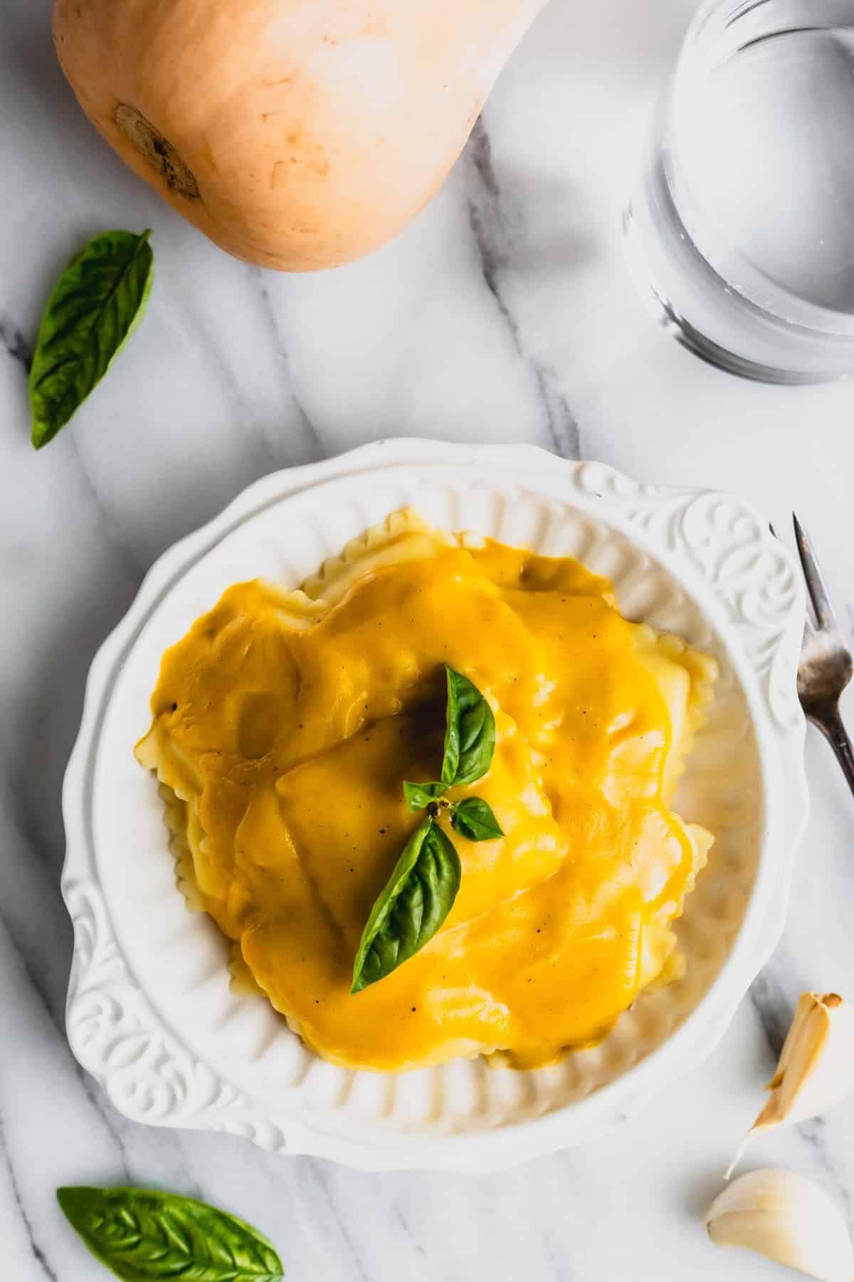 Overhead view of ravioli covered with butternut squash sauce in a white bowl with basil, garlic, butternut squash, a glass of water and a fork around it.