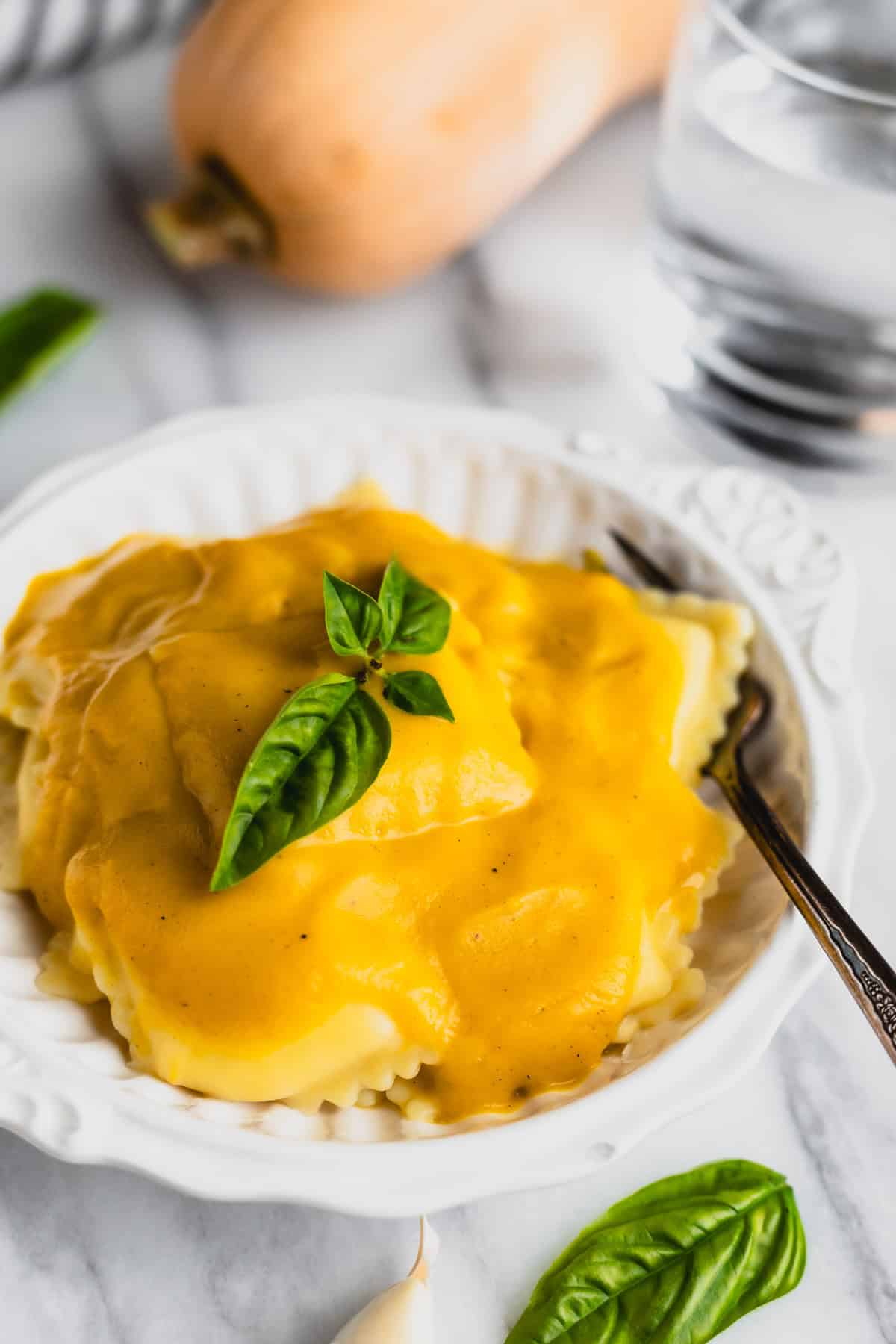 Ravioli covered in butternut squash sauce in a white bowl with a glass of water, basil and a whole butternut squash in the background.