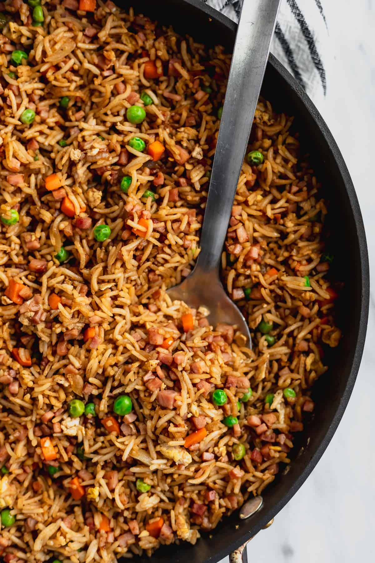 Close up of part of a skillet of ham fried rice with a large, silver spoon in it.