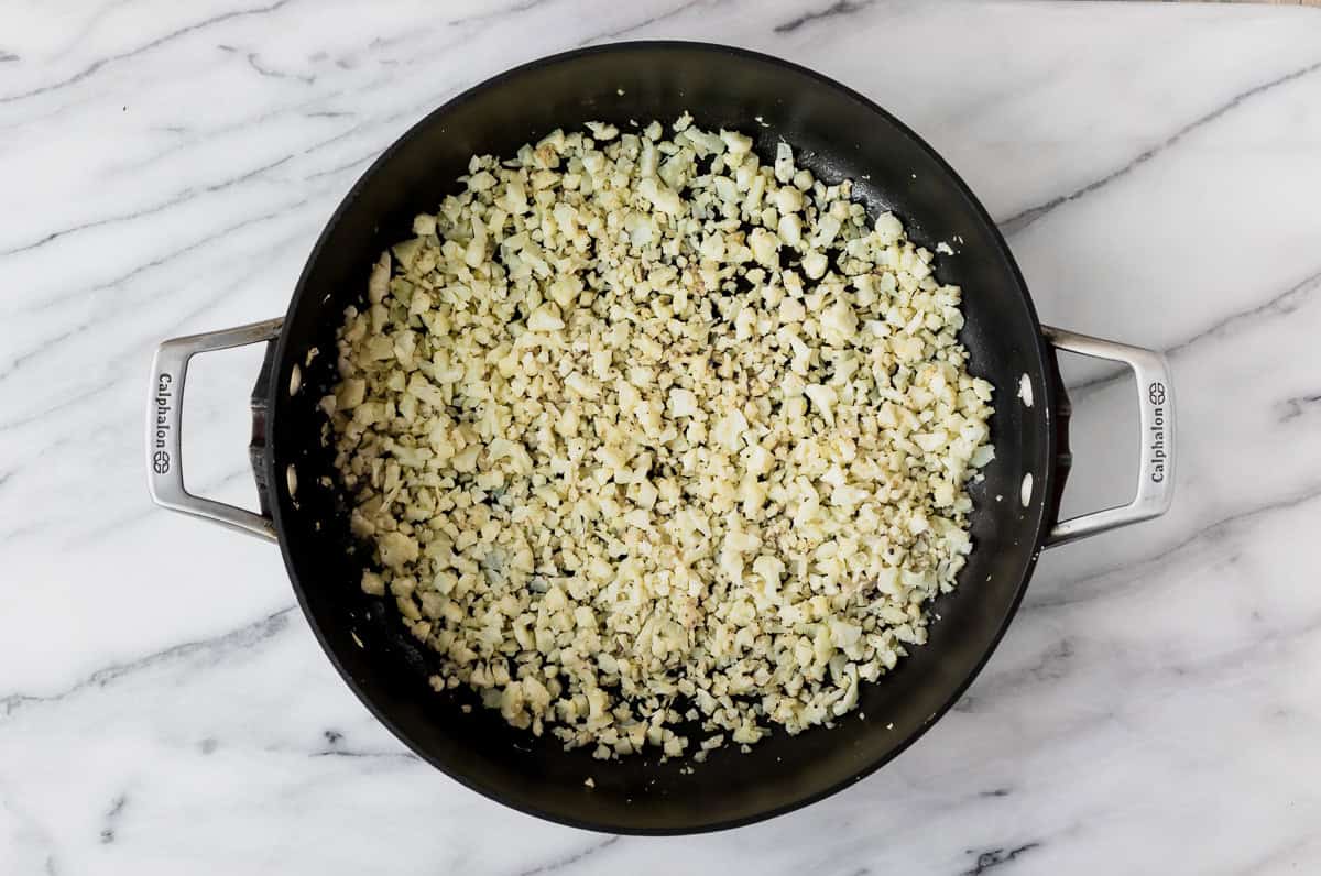 Cooked cauliflower rice in a black skillet over a marble background.
