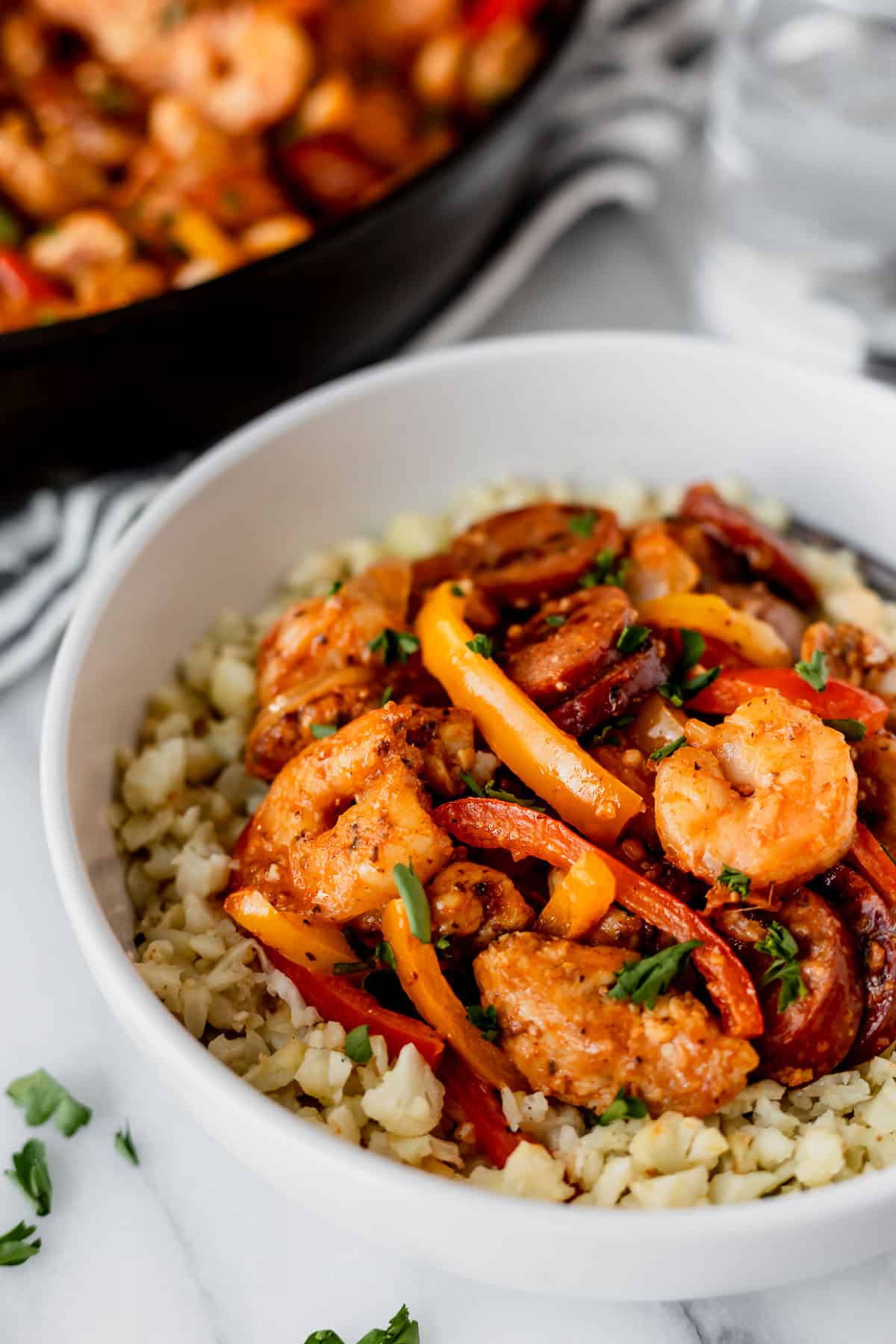 A white bowl filled with cauliflower rice and jambalaya with the black skillet in the background.