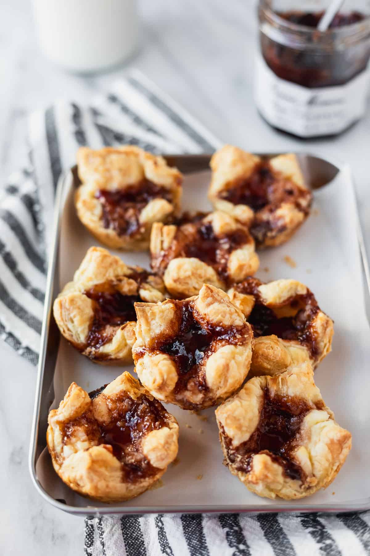 Sugar Plum Cheese Danishes on a parchment paper lined baking sheet over a striped towel with a glass of milk and jar of plum preserves in the background.