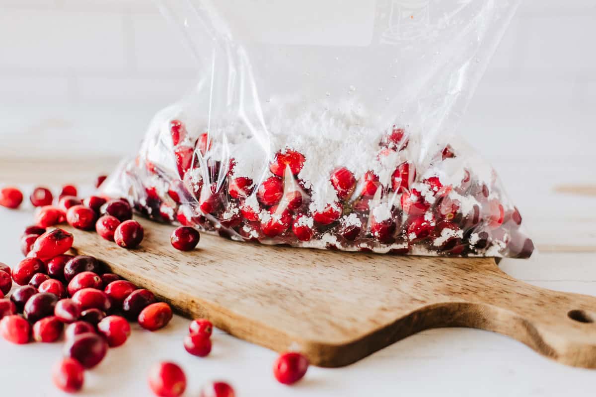 Cranberries and powdered sugar in a ziplok bag on a wood board with more cranberries around it.