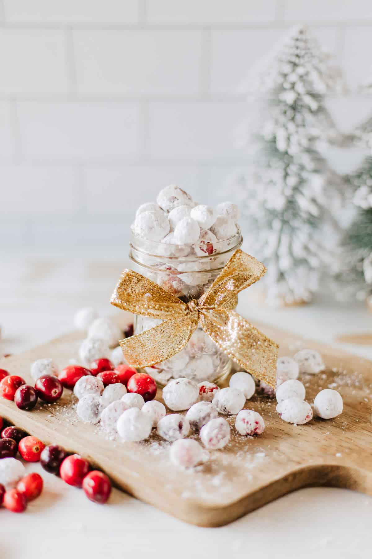 A jar of candied cranberries surround by more candied and plain cranberries on a wood board with pine trees in the background.