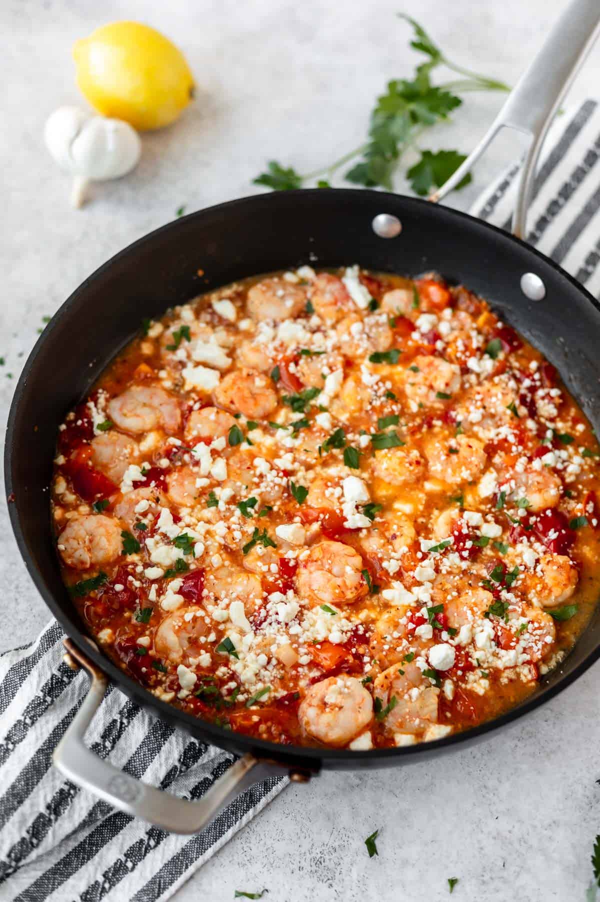 A skillet full of Greek shrimp with tomatoes and feta cheese over a striped towel, lemon and garlic bulb on a light background.
