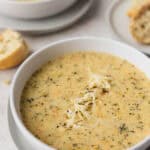 A close up of Instant Pot Broccoli Cheese Soup in a white bowl on a white plate with a second bowl and bread on the background with text overlay.