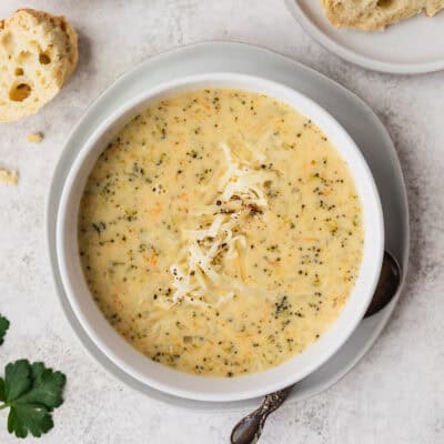 Overhead view of a bowl of Instant Pot Broccoli Cheese Soup in a white bowl over a white plate with a spoon, parsley and crusty bread partially showing around it.
