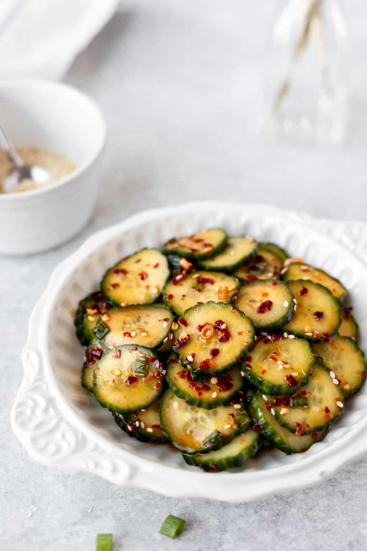 Korean cucumber salad with red pepper flakes in a white bowl over a light background with a white bowl and glass bottle in the background.
