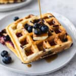 Syrup being poured over two blueberry waffles that are stacked on a white plate with a second plate partially showing in the background.