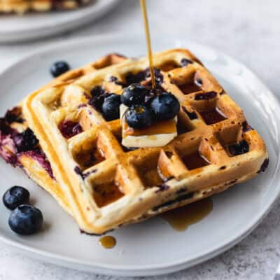 Syrup being poured over two blueberry waffles that are stacked on a white plate with a second plate partially showing in the background.