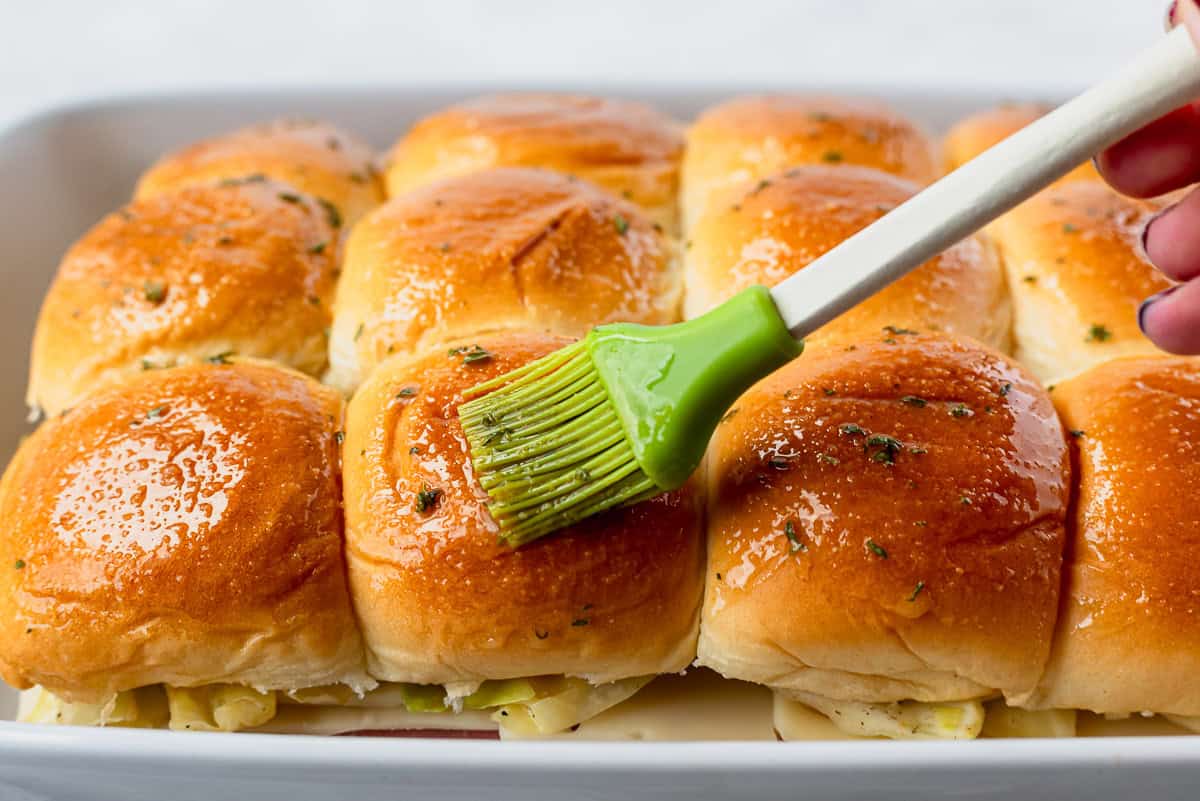 Close up of melted butter and herbs being brushed over the tops of Hawaiian rolls.
